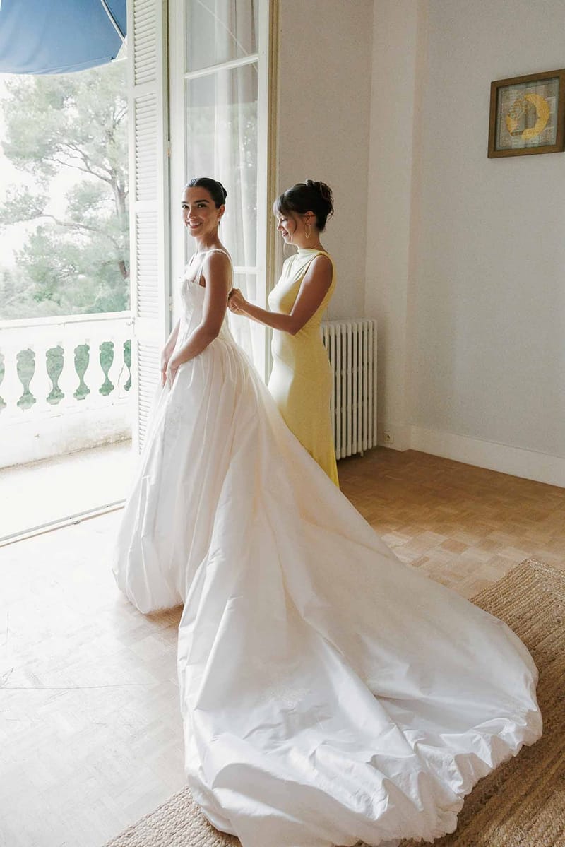 Bridesmaid helping the bride into her gown in a sunlit bedroom at Domaine Rocabella, French Riviera