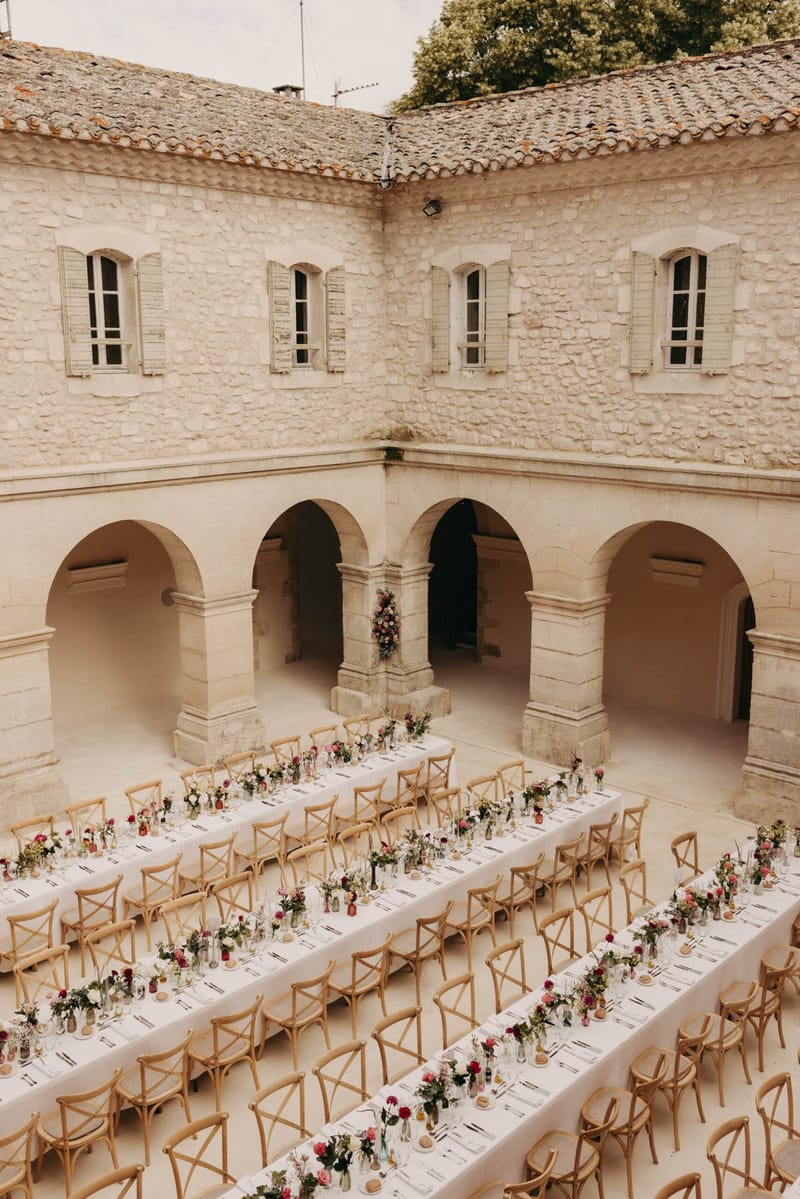 Aerial banquet tables with fuchsia and burgundy bud vase runners under stone arcade with terracotta roof and shutters