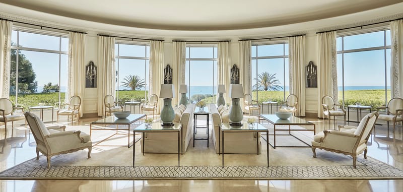 Curved salon with arched windows, ivory drapes, cream Louis XVI seating, and palm trees and sea view beyond
