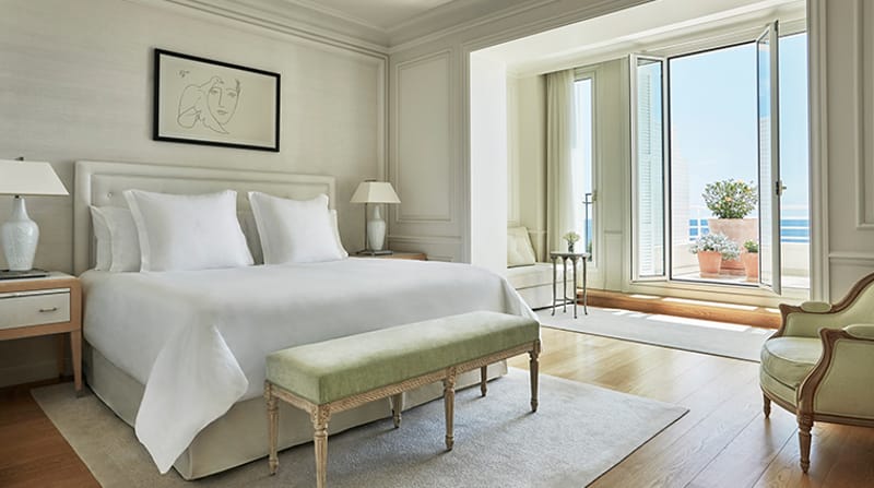 Hotel bedroom suite with king bed, sage green bench, armchair, and French doors opening to sea view terrace