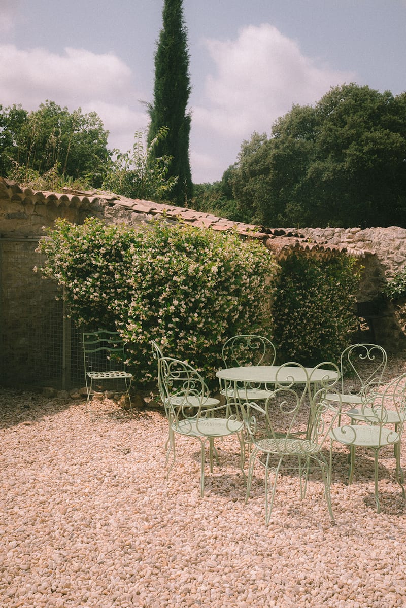 French country courtyard with sage green wrought iron garden furniture on gravel beside a stone building with climbing flo...