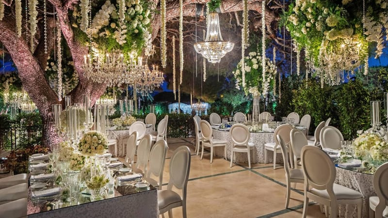 Outdoor evening reception setup with crystal chandeliers hanging from trees, white rose centerpieces and Louis XVI chairs