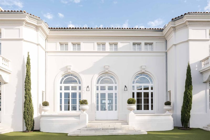 White rendered villa with three arched doors, columnar cypress trees, and topiary urn planters on steps