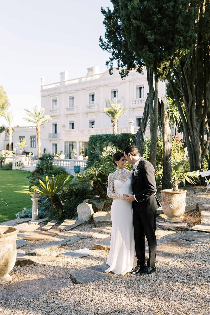 Bride in lace bodice gown and groom in charcoal suit touching foreheads on gravel path before white Belle Epoque villa