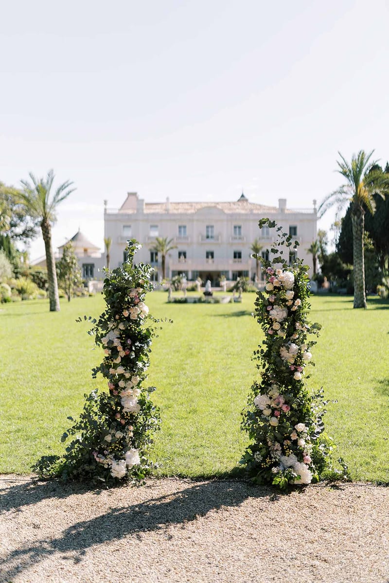 Two greenery-and-rose floral columns frame gravel path leading to neoclassical white chateau on lawn