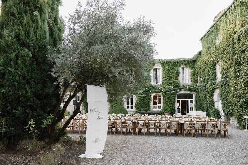 Long tables with white linens and welcome banner before ivy-covered stone manor with string lights on gravel courtyard