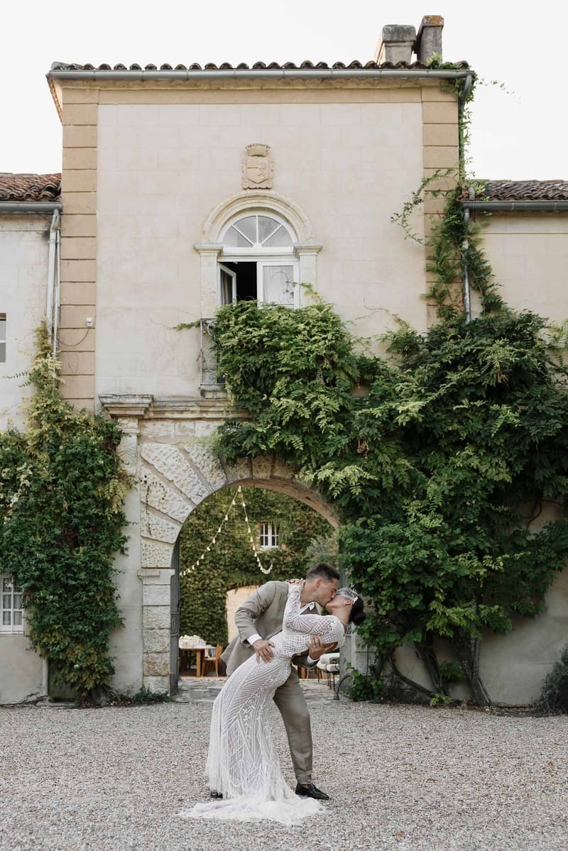 Groom dipping bride in beaded fringe gown before vine-covered chateau archway with fairy-lit reception