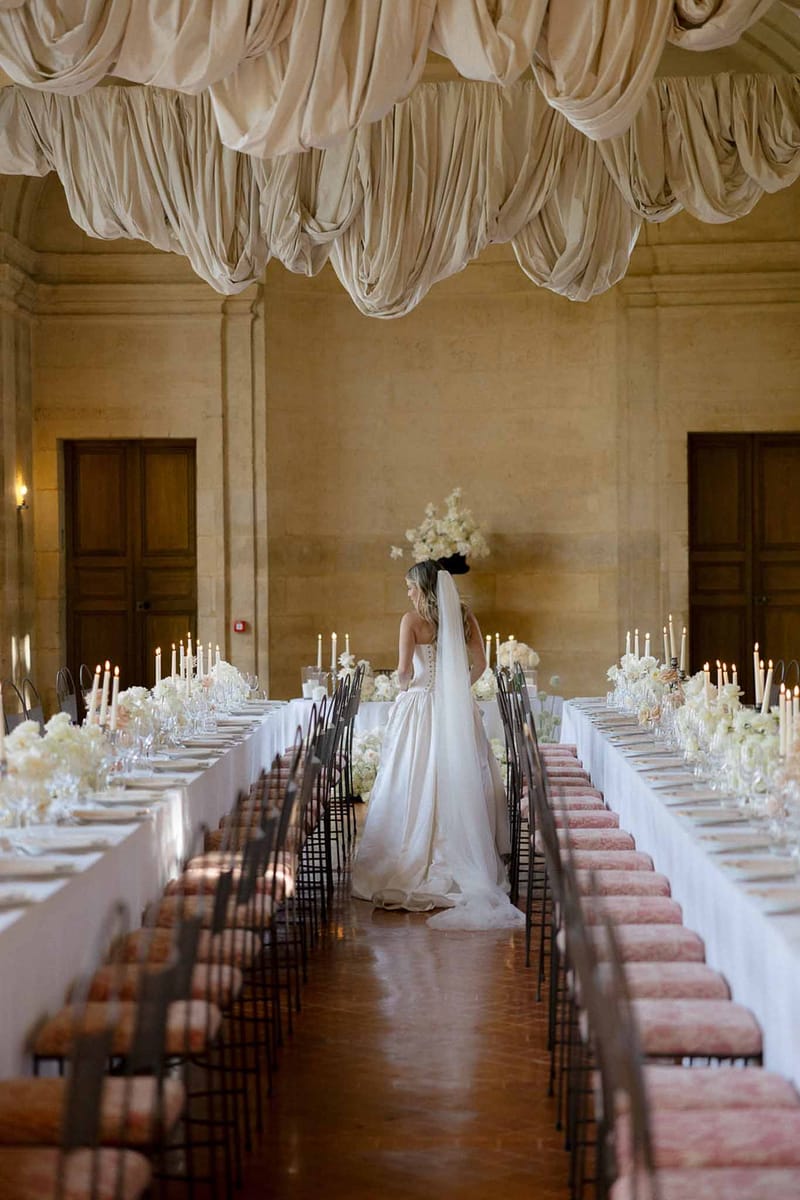 Bride in ballgown standing between two long banquet tables in a grand hall with draped fabric ceiling
