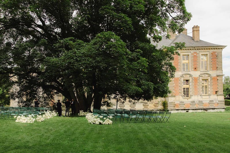Teal folding chairs on lawn with white hydrangea aisle clusters before brick-and-stone chateau