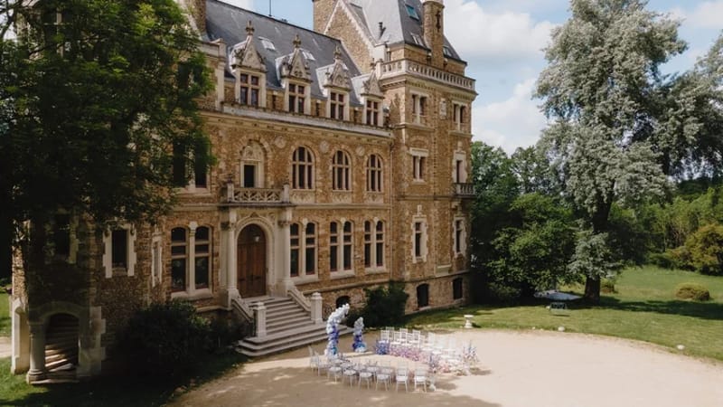 Aerial Gothic-Revival chateau with white ceremony chairs and blue-white floral arch on gravel forecourt
