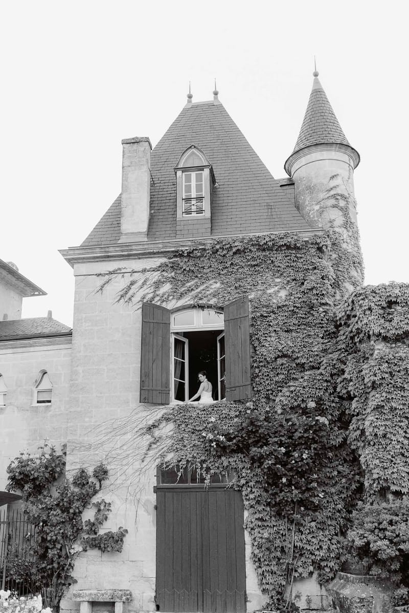 Black-and-white shot of bride at open chateau window with ivy-covered stone facade and slate turret roof