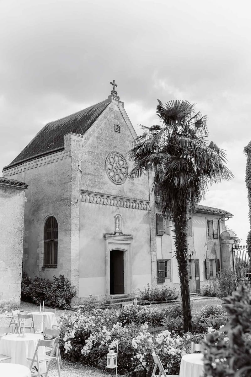 Romanesque estate chapel with rose window and palm tree beside cocktail tables on garden in B&W