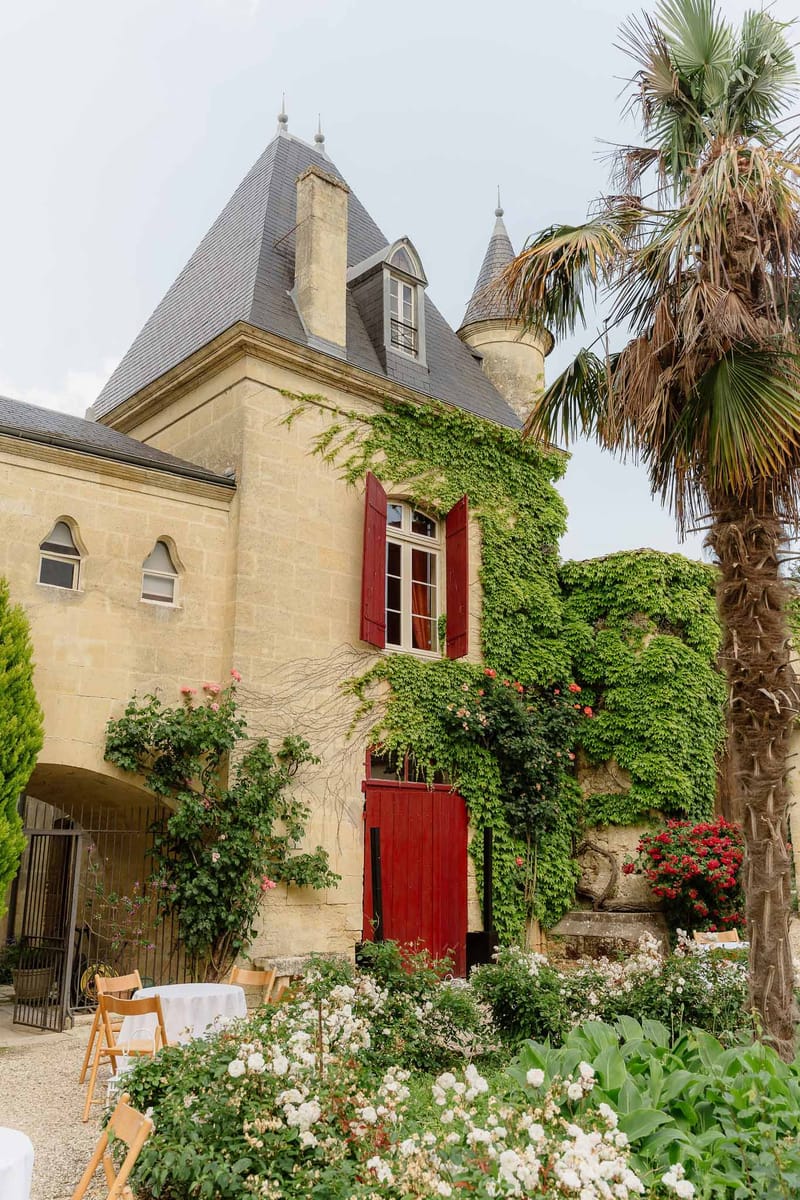 Honey-stone chateau with slate turrets, red shutters, and ivy beside white-clothed courtyard tables