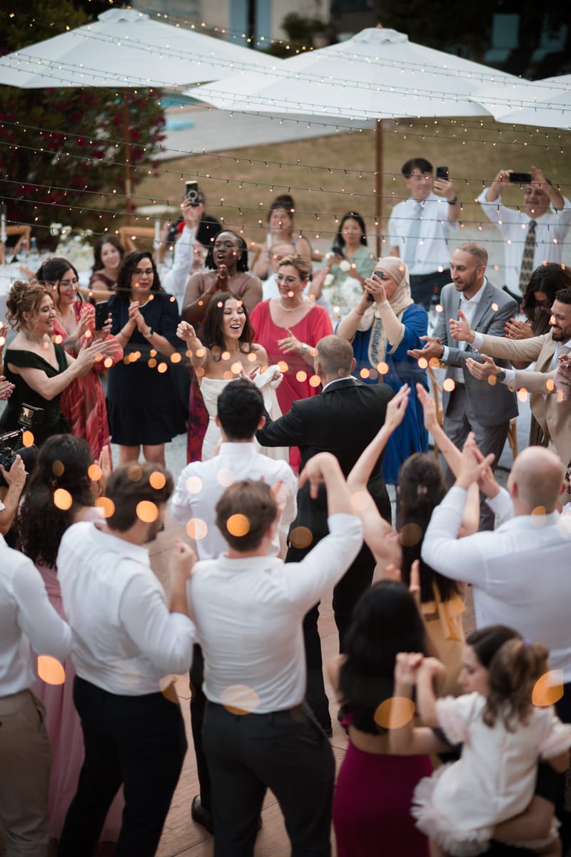 Bride and groom dancing in center of guest circle on outdoor terrace with fairy lights and white umbrellas at evening rece...