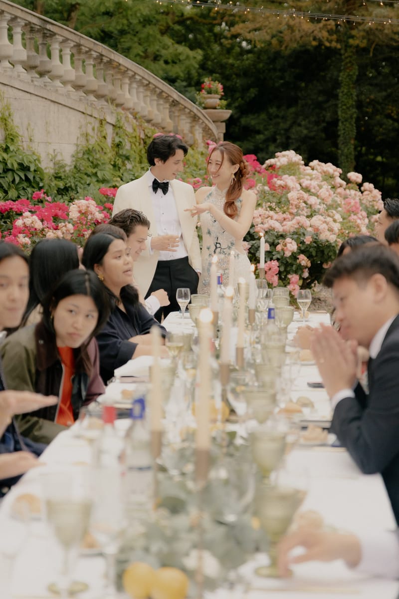 Bride in floral cheongsam and groom greeting guests at outdoor garden reception with fairy lights