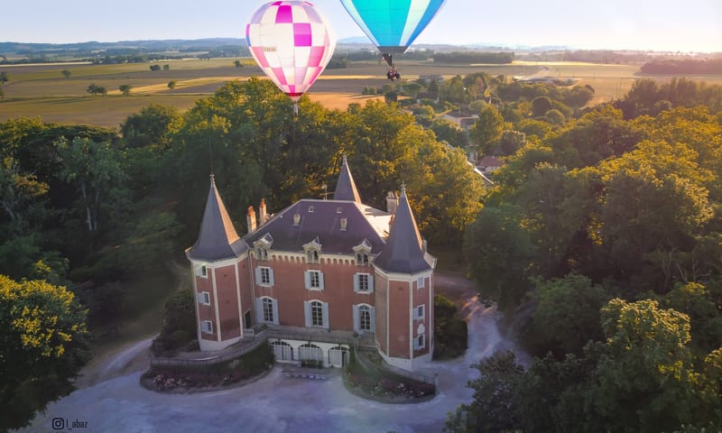 Aerial red-brick chateau with conical towers and orangery as two hot air balloons fly overhead at golden hour