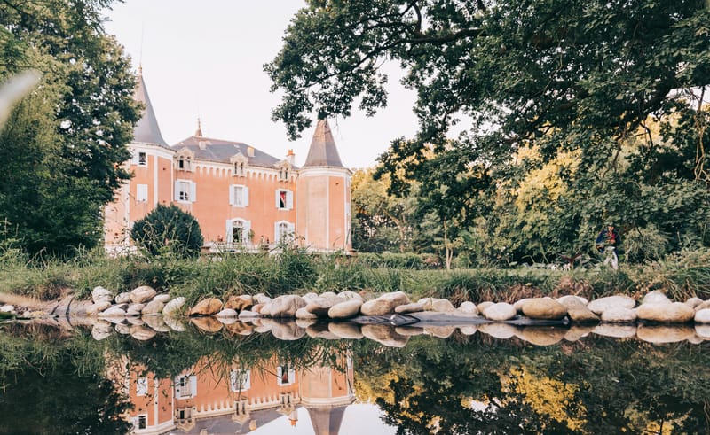 Terracotta-pink chateau with conical slate towers reflected in still ornamental pond bordered by white stones