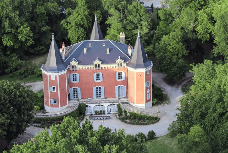 Aerial view of terracotta chateau with corner turrets pale blue shutters veranda and mature woodland