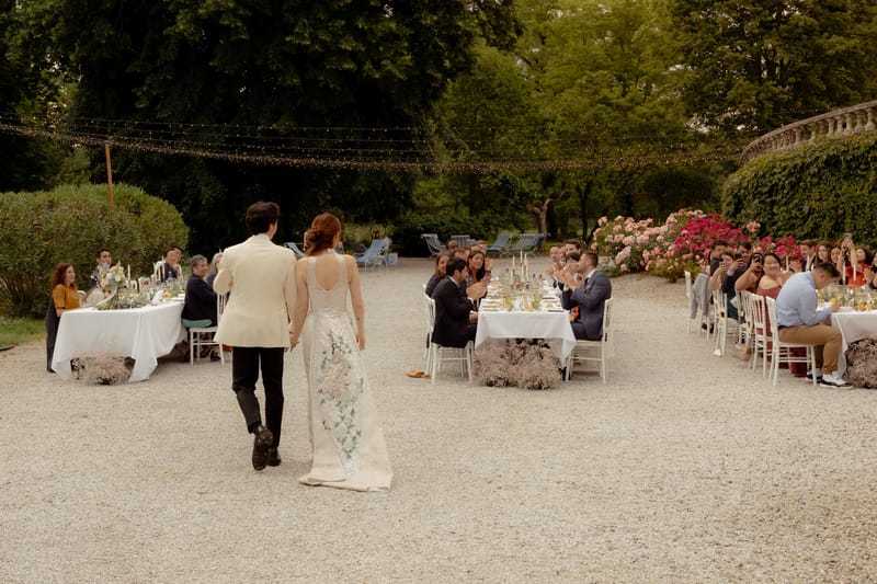 Bride and groom entering garden reception between banquet tables with fairy lights and thirty applauding guests