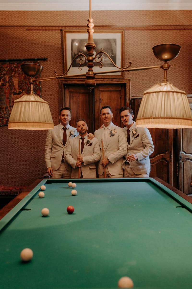 Groom and three groomsmen in cream linen suits with burgundy ties posing behind a snooker table in a chateau billiards room