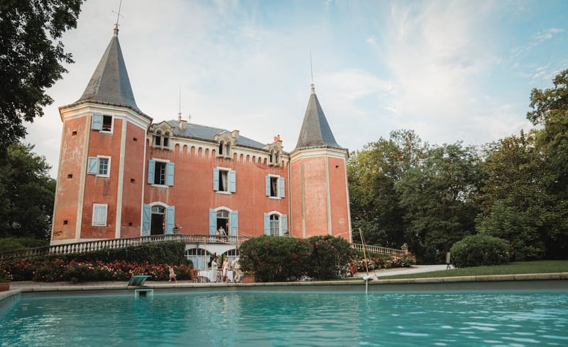 Red brick chateau with conical towers, blue shutters, and turquoise pool at golden hour with guests