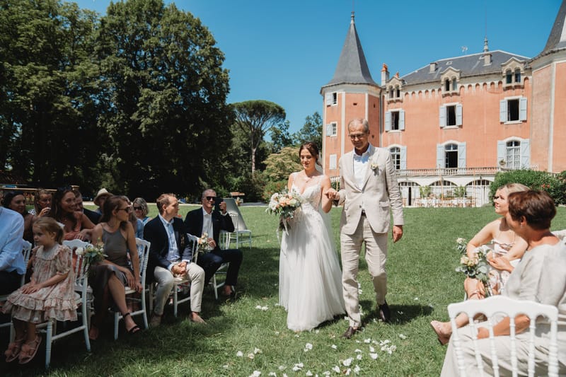 Bride walked down aisle by father at pink chateau with turret, white Chiavari chairs, and scattered petals