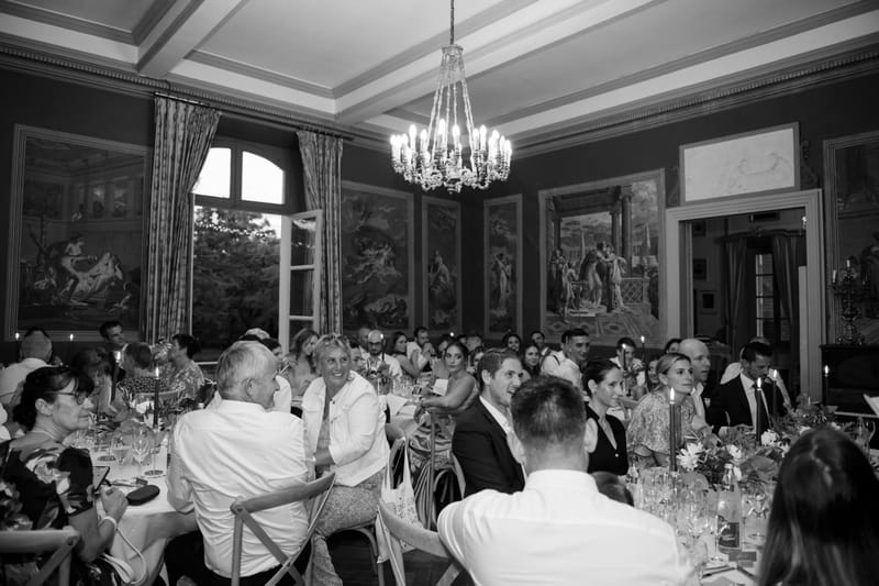 Grand ballroom reception with 50-plus guests at long tables beneath crystal chandelier and classical murals in B&W