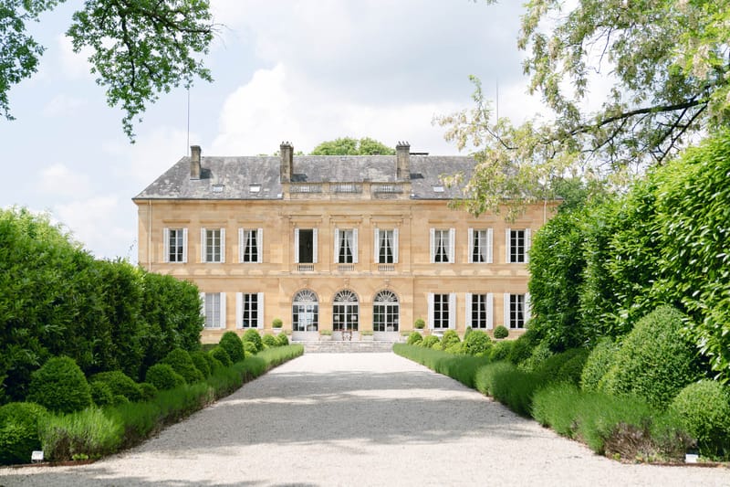 Golden limestone chateau facade with white shutters, box topiary-lined gravel driveway, and lavender borders