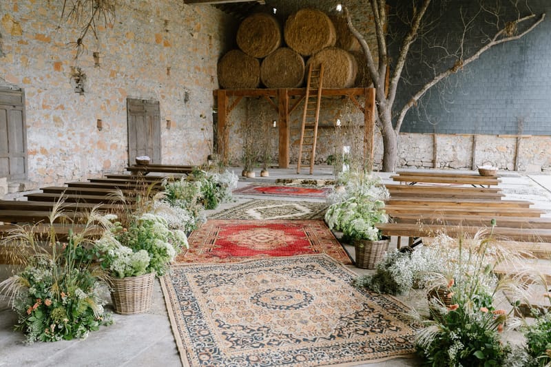 Rustic stone barn ceremony setup with vintage rugs lining the aisle, hay bale altar, and meadow-style floral baskets