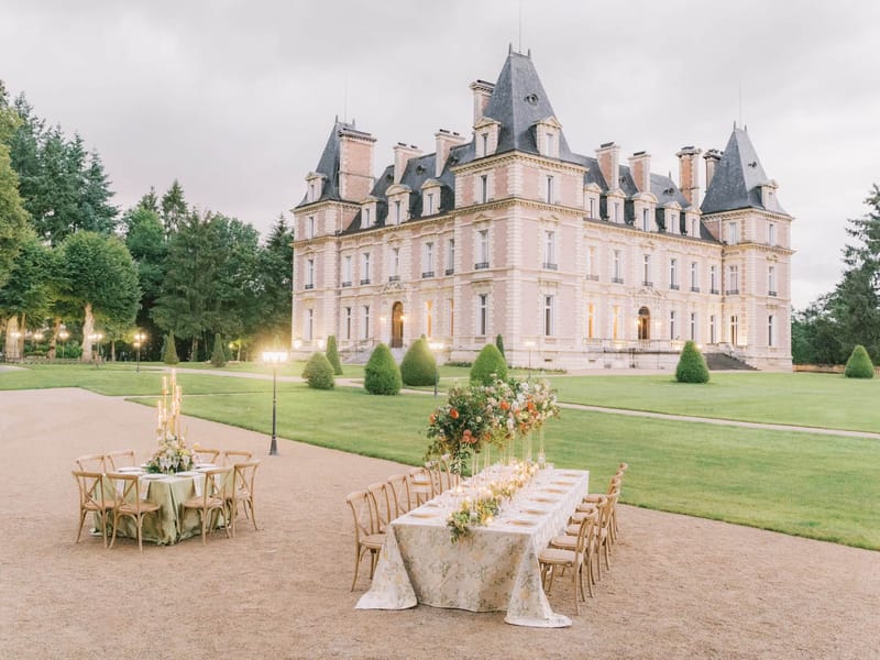 Outdoor reception tables with floral centerpieces and gold chargers on chateau gravel forecourt at dusk