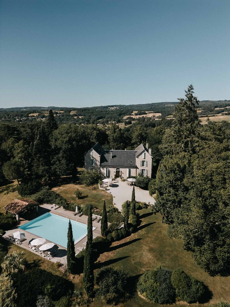 Aerial drone view of French stone manor with swimming pool, cypress trees, and rolling countryside