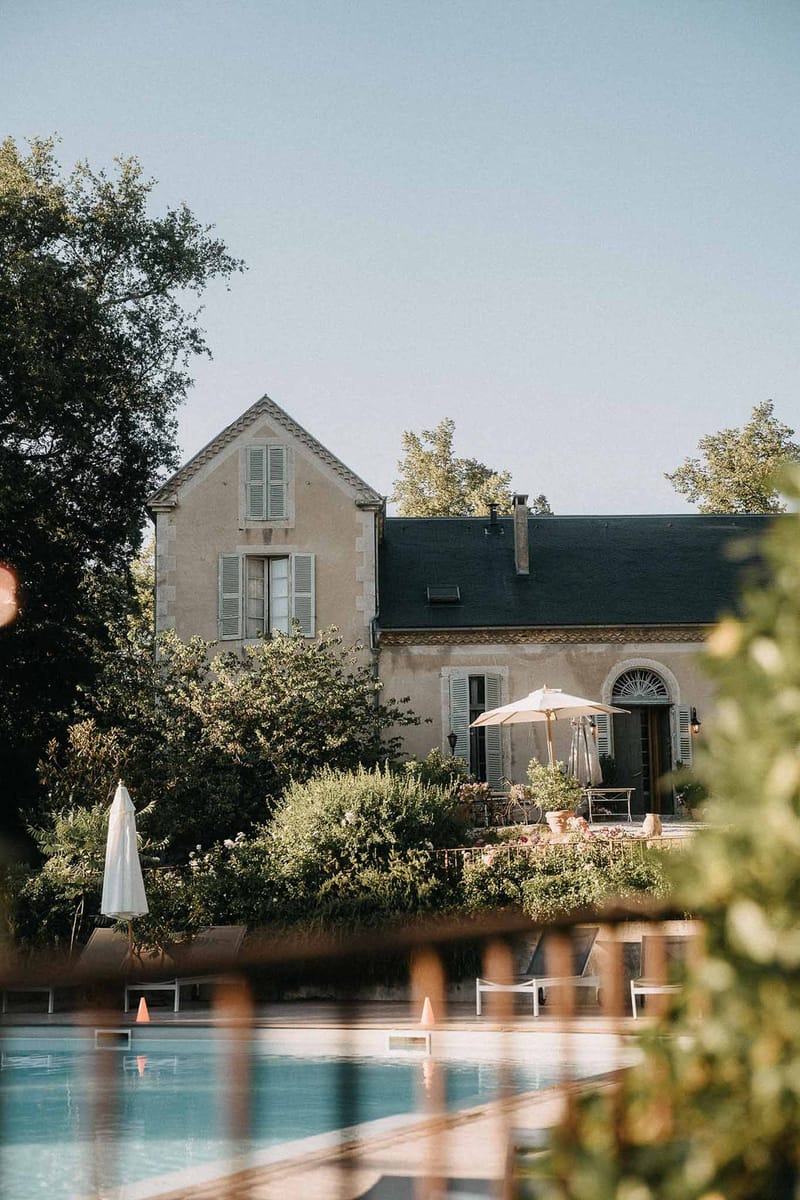 French manor house viewed across swimming pool with cream parasol, white loungers, and slate roof