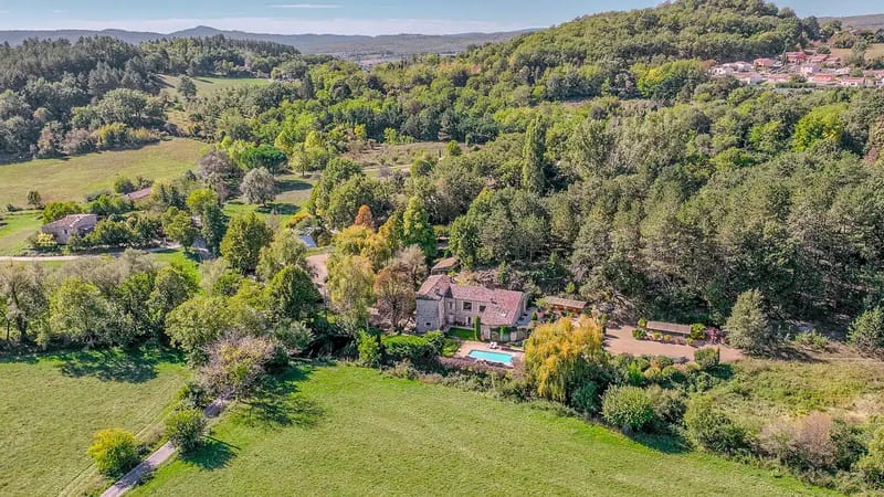 Aerial view of stone country house with pool, gravel courtyard, and wooded valley landscape
