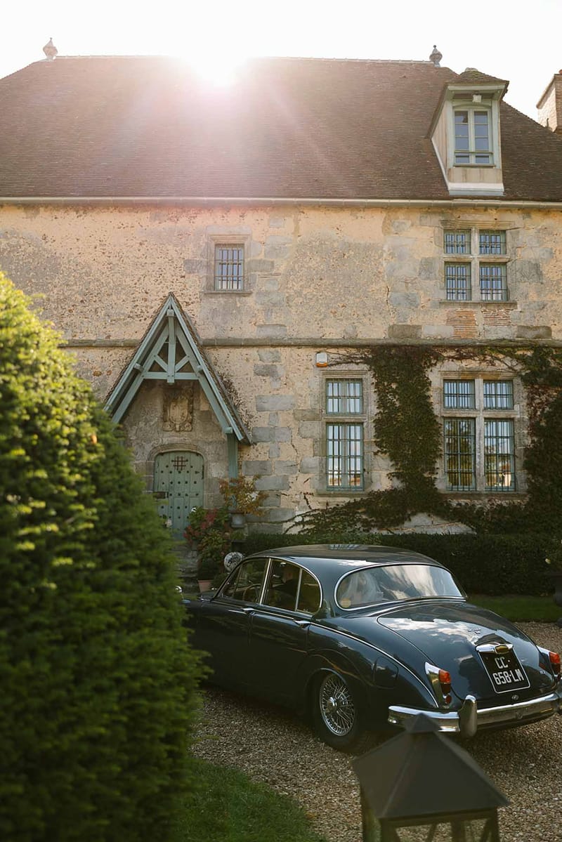 French chateau with limestone walls and slate roof at golden hour with navy vintage Jaguar parked on gravel driveway