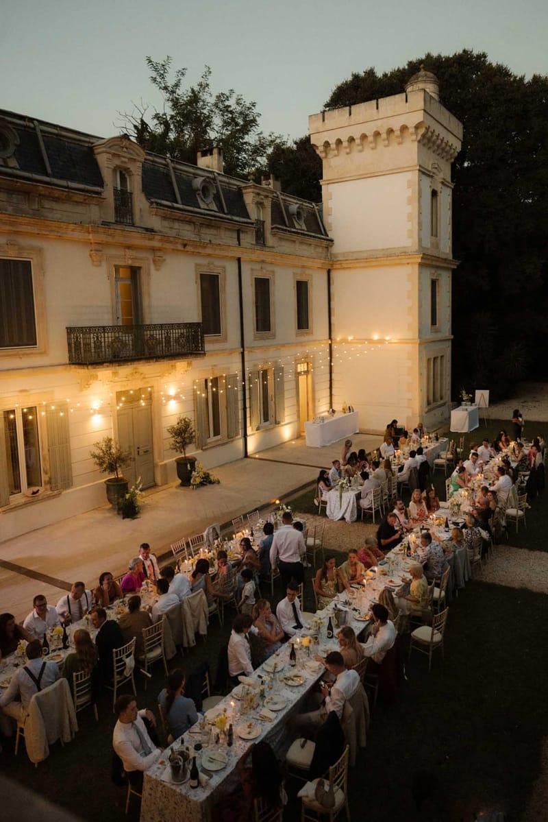 Elevated view of outdoor chateau courtyard wedding reception at dusk with fairy lights and guests at long banquet tables