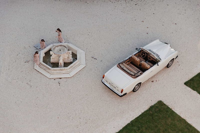 Aerial view of three flower girls in blush tulle dresses gathered around octagonal fountain beside vintage cream convertible