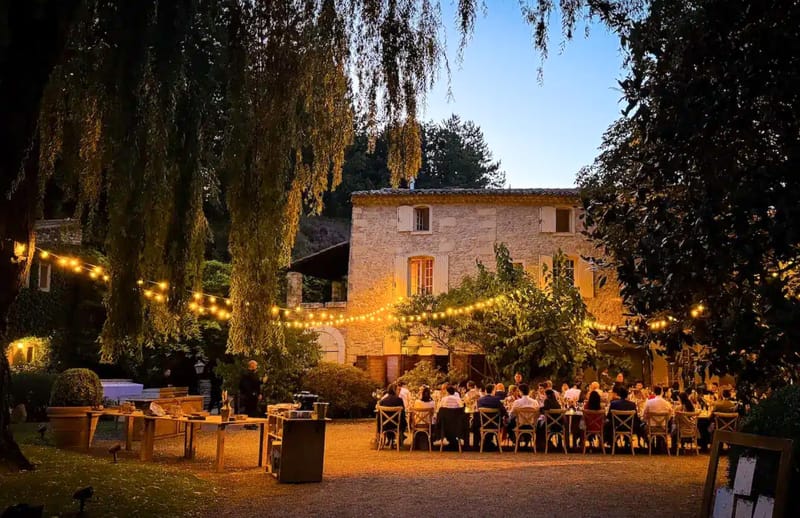 Dusk reception at stone mas courtyard with cross-back chairs and amber string lights under willows