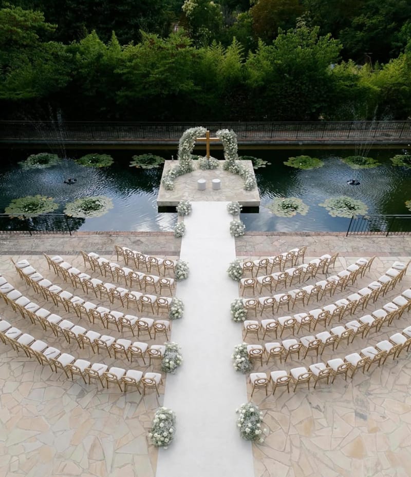 Aerial view of outdoor wedding ceremony setup with gold chairs and white floral arch over ornamental pond