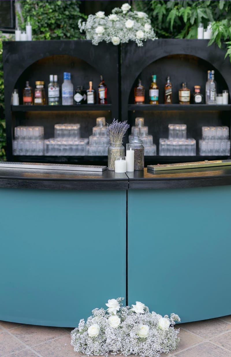 Teal curved cocktail bar with white rose and gypsophila arrangements against green foliage backdrop