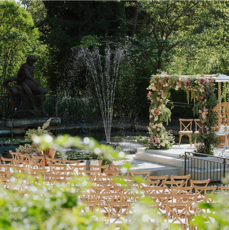 Ceremony setup with square floral arch in blush and coral tones, peacock chairs, and lily pond with fountain