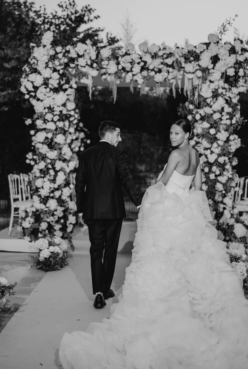 Bride and groom walk from square floral arch at evening outdoor ceremony with Chiavari chairs in B&W