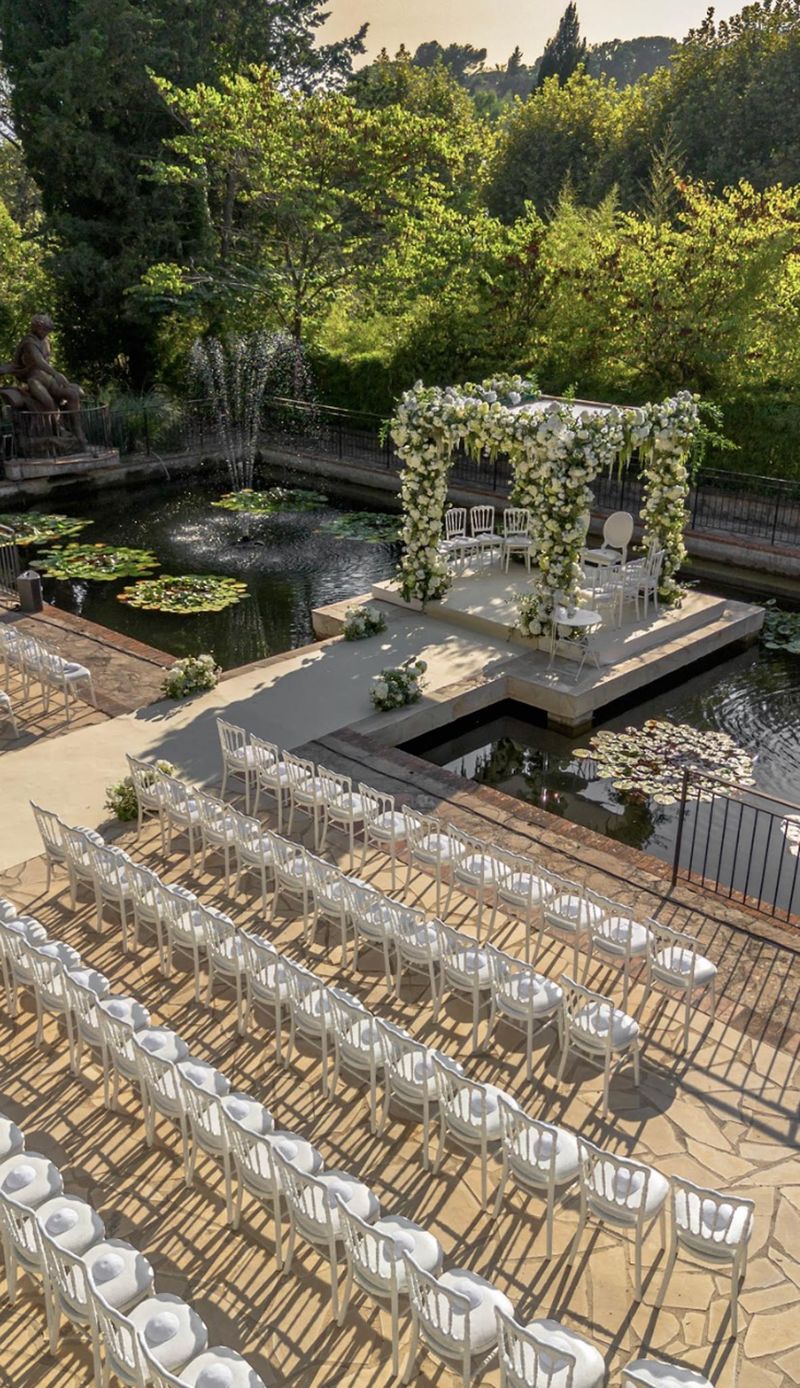 Aerial view of outdoor ceremony setup over lily pond with square floral arch and white chair rows