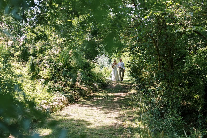 Bride and groom walking arm in arm along wooded garden path framed by overhanging tree canopy