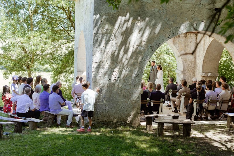 Wedding ceremony beneath historic stone arcade with couple exchanging vows and 40-50 seated guests
