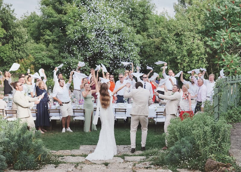 Bride and groom enter outdoor garden reception as guests cheer, wave napkins, and toss white petals along banquet table