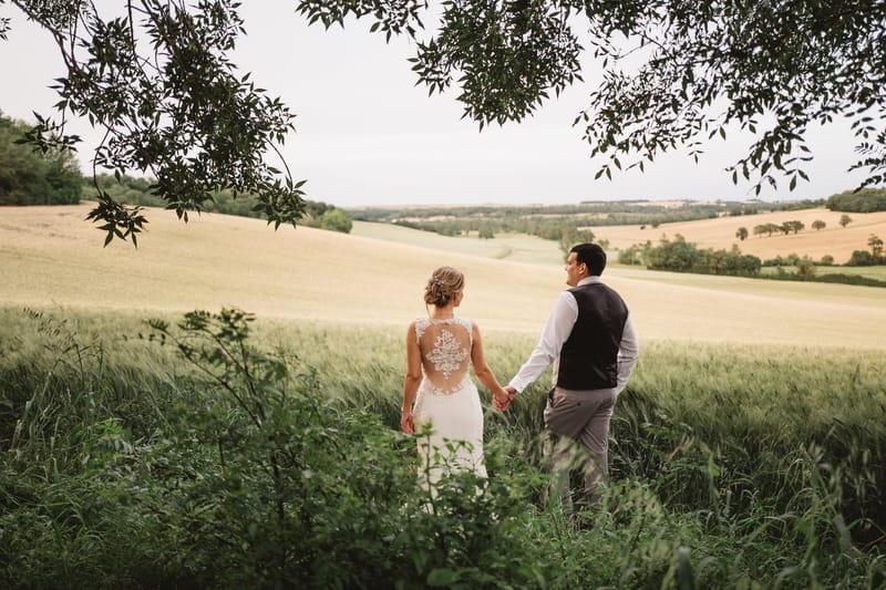 Bride in ivory lace-back gown and groom in navy waistcoat walking hand in hand through wild grass in the French countryside