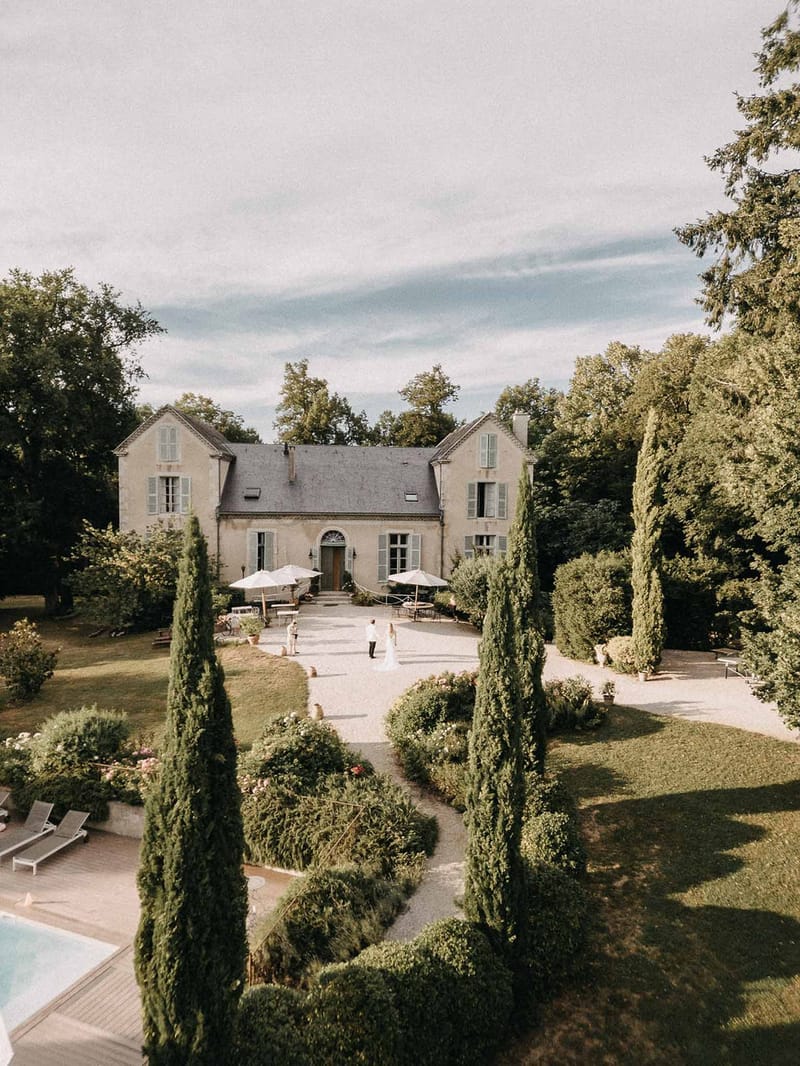 Aerial view of stone manor with cypress-lined drive, formal gardens, pool, and couple on gravel courtyard