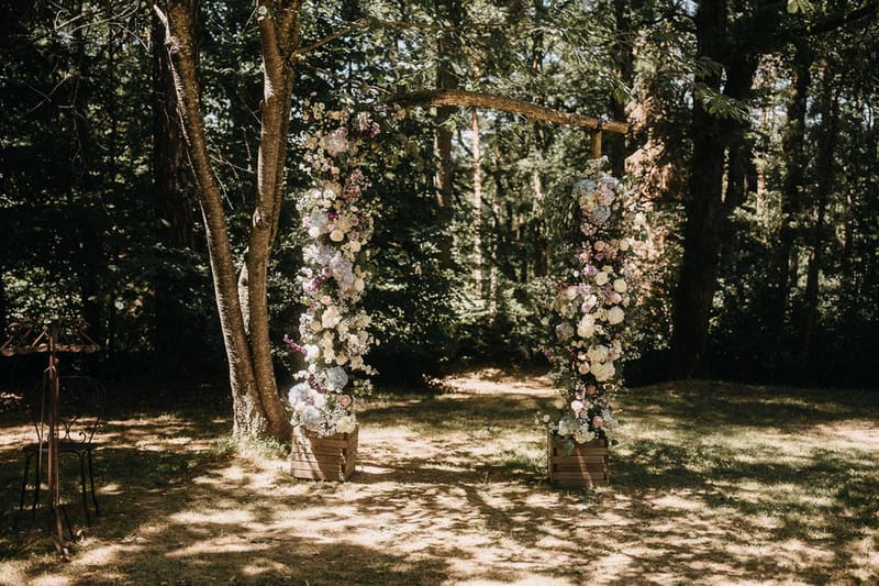 Rustic wooden ceremony arch decorated with ivory hydrangeas, blush roses, and mauve blooms in a sunlit forest clearing
