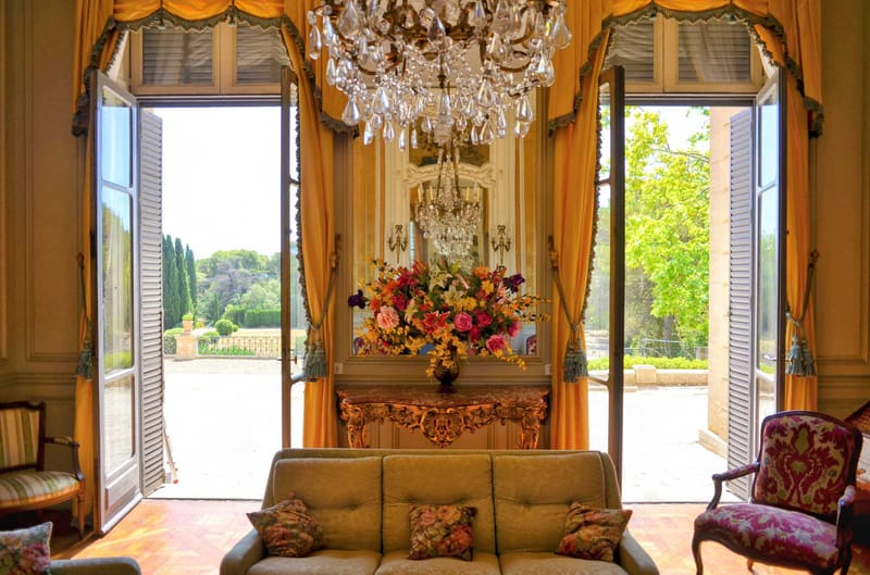 Grand chateau salon with crystal chandelier, gold drapes, Louis XV furniture, and French doors open to formal garden
