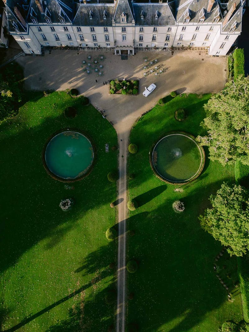 Aerial top-down view of a cream stone chateau with symmetrical fountains, gravel driveway, and vintage wedding car